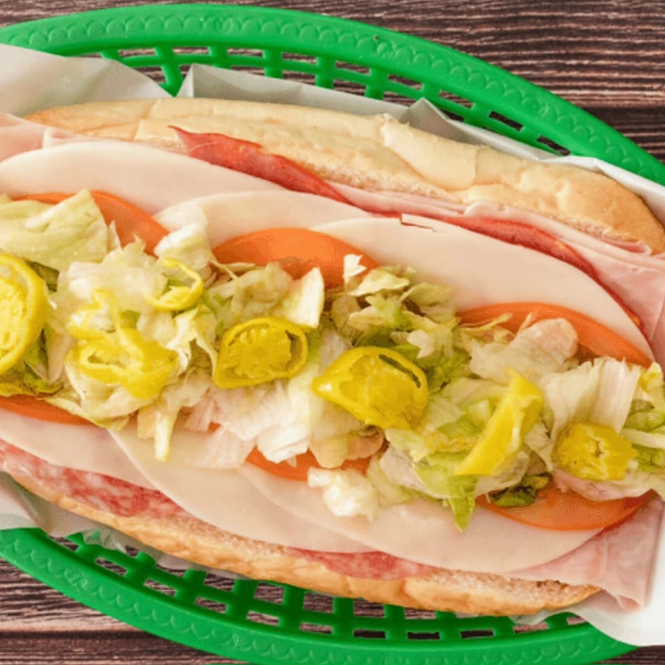 A large deli sub sandwich with various meats, cheese, lettuce, tomatoes, and banana peppers in a green basket.
