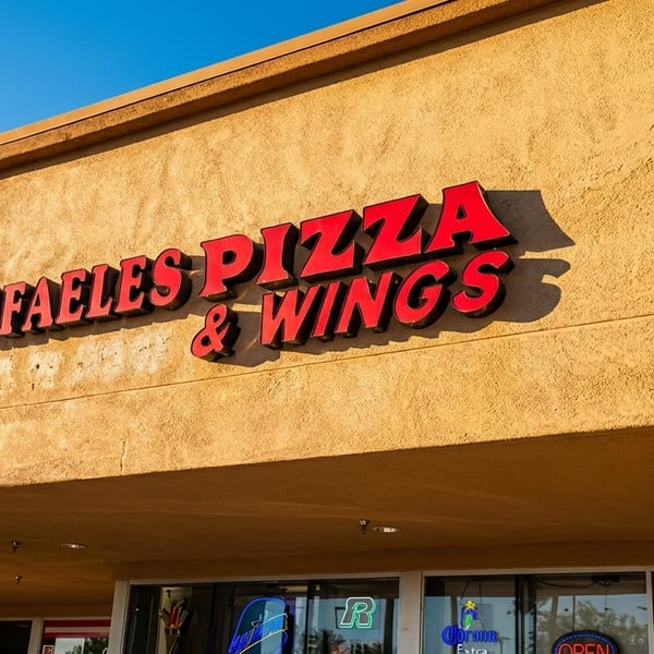 Raffaeles Pizza & Wings sign with red letters on a textured tan building under a blue sky.