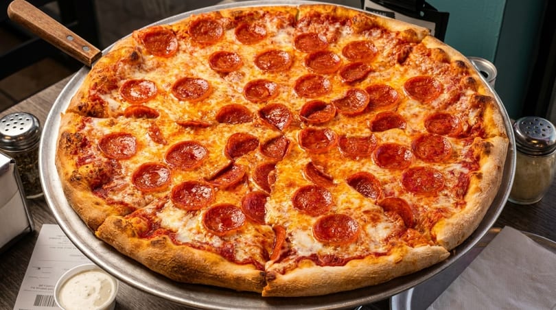 A large, hot pepperoni pizza with a slice removed, on a metal tray with shakers and ranch.