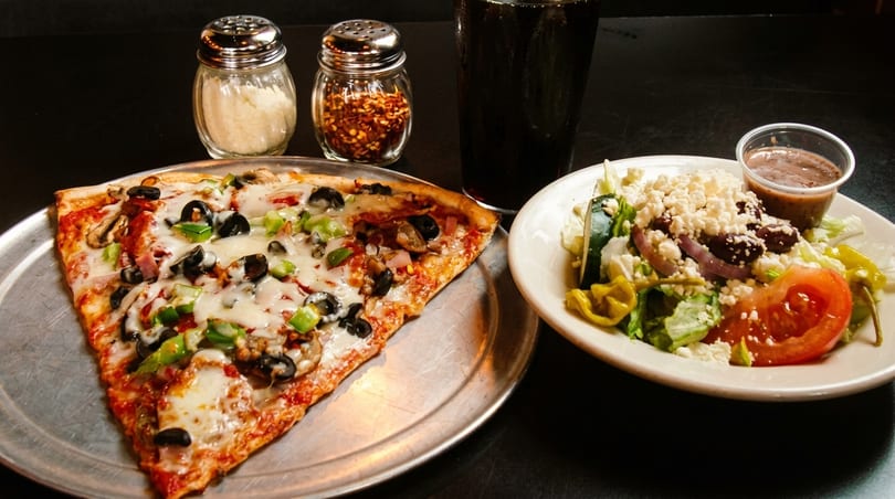 A slice of pizza with a side salad, drink, and shakers of cheese and red pepper flakes.