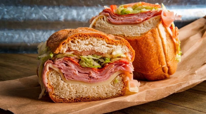 Two large, sliced Italian subs with layers of meat, cheese, lettuce, and tomato on brown paper.